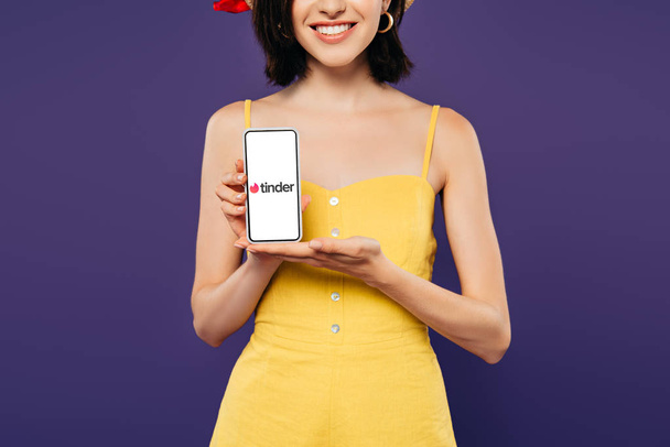 KYIV, UKRAINE - JULY 3, 2019: cropped view of smiling girl holding smartphone with tinder app isolated on purple  - Fotografie, Obrázek