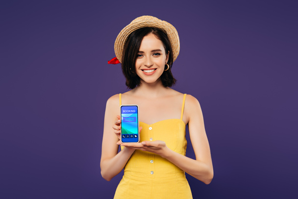 улыбающаяся красивая девушка в соломенной шляпе, держащая смартфон с приложением бронирования, изолированным на фиолетовый
  - Фото, изображение