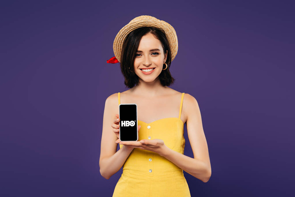 KYIV, UKRAINE - JULY 3, 2019: smiling pretty girl in straw hat holding smartphone with HBO app isolated on purple  - Fotografie, Obrázek