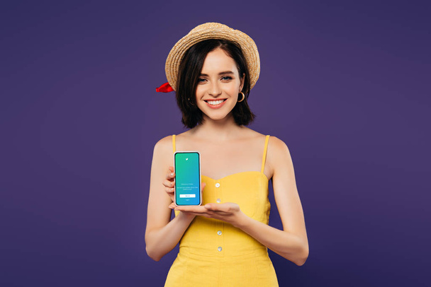 KYIV, UKRAINE - JULY 3, 2019: smiling pretty girl in straw hat holding smartphone with twitter app isolated on purple  - Fotó, kép