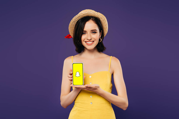 KYIV, UKRAINE - JULY 3, 2019: smiling pretty girl in straw hat holding smartphone with snapchat app isolated on purple  - Foto, afbeelding