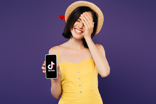 KYIV, UKRAINE - JULY 3, 2019: smiling pretty girl in straw hat with hand on face showing smartphone with tiktok app isolated on purple  - Fotografie, Obrázek