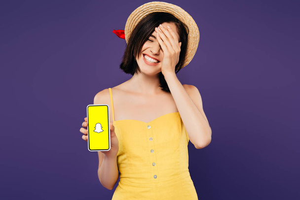 Kijów, Ukraina-3 lipca 2019: uśmiechnięta Ładna dziewczyna w słomy kapelusz z ręki na twarzy pokazując smartphone z Snapchat aplikacji na białym tle  - Zdjęcie, obraz