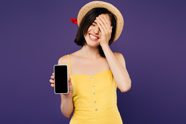улыбается красивая девушка в соломенной шляпе с рукой на лице показывая смартфон с пустым экраном изолированы на фиолетовый
  - Фото, изображение