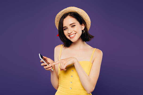 smiling pretty girl in straw hat holding smartphone isolated on purple  - Photo, Image
