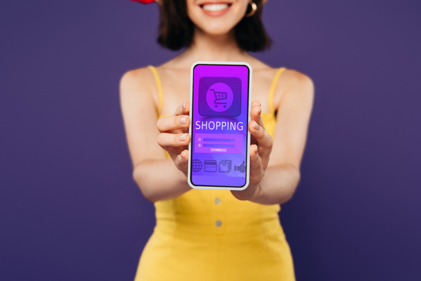 selective focus of smiling girl presenting smartphone with online shopping app isolated on purple  - Photo, Image