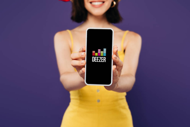 Kyjev, Ukrajina-3. července 2019: selektivní zaměření usměvavé dívky prezentuje smartphone s Deezer aplikací izolované na purpurové  - Fotografie, Obrázek