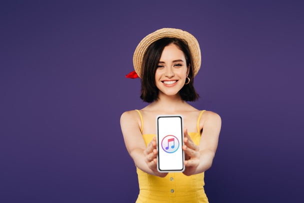 Kiev, Oekraïne-juli 3, 2019: selectieve focus van glimlachend mooi meisje in stro hoed presenteren van smartphone met iTunes-App geïsoleerd op paars  - Foto, afbeelding