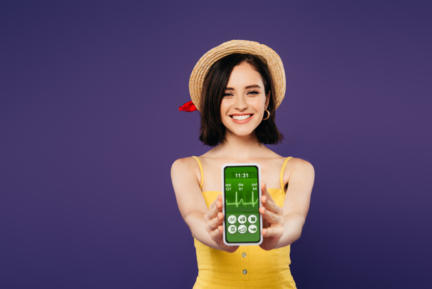 selective focus of smiling pretty girl in straw hat presenting smartphone with healthcare app isolated on purple  - Photo, Image