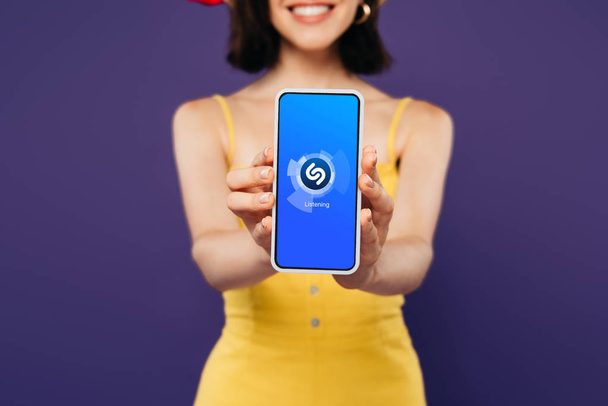 KYIV, UCRANIA - 3 de julio de 2019: enfoque selectivo de la chica sonriente que presenta el teléfono inteligente con la aplicación shazam aislada en púrpura
  - Foto, Imagen