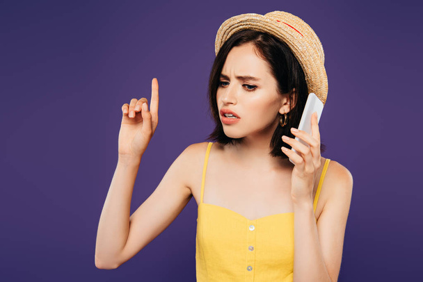 sad pretty girl in straw hat pointing with finger and talking on smartphone isolated on purple  - Photo, Image