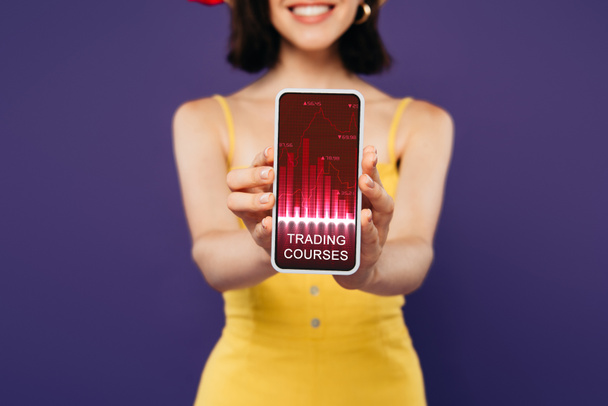 selective focus of smiling girl presenting smartphone with trading courses app isolated on purple  - Photo, Image