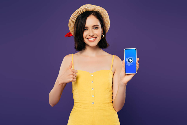 KYIV, UKRAINE - JULY 3, 2019: smiling pretty girl in straw hat showing thumb up while holding smartphone with shazam app isolated on purple  - Foto, imagen