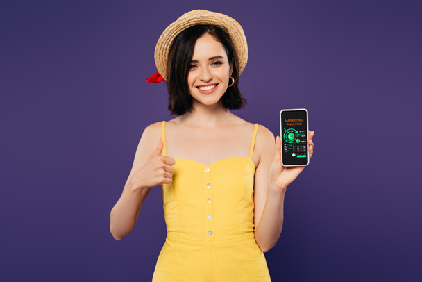 улыбающаяся красивая девушка в соломенной шляпе показывает большой палец вверх, держа смартфон с приложением маркетингового анализа изолированы на фиолетовый
  - Фото, изображение