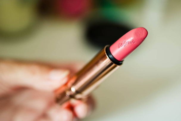 Lápiz labial rosa con inscripción ser valiente en la mano de las mujeres. Fondo abstracto borroso. Concepto de moda
. - Foto, imagen