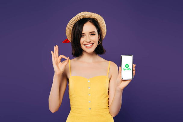 KYIV, UKRAINE - JULY 3, 2019: smiling pretty girl in straw hat holding smartphone with spotify app and showing ok sign isolated on purple  - Фото, изображение