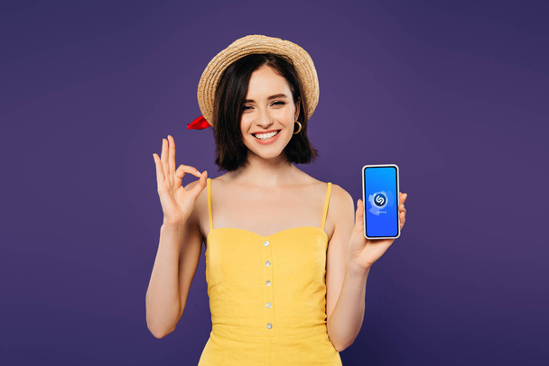 KYIV, UCRANIA - 3 DE JULIO DE 2019: niña bonita sonriente con sombrero de paja sosteniendo el teléfono inteligente con la aplicación Shazam y mostrando un signo aceptable aislado en púrpura
  - Foto, imagen