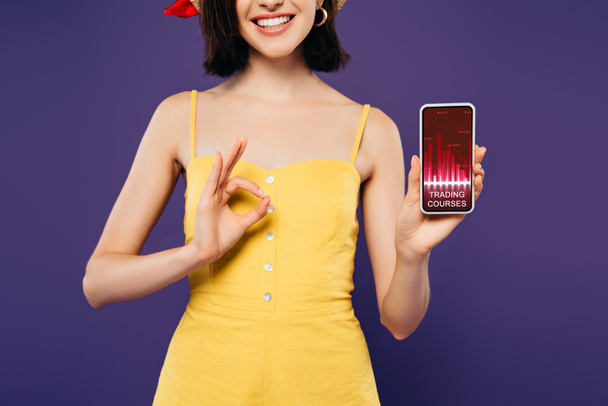 περικομμένη θέα του χαμογελαστή κορίτσι στο κράτημα smartphone με τα μαθήματα συναλλαγών app και δείχνοντας OK πρόσημο απομονωθεί σε μωβ  - Φωτογραφία, εικόνα