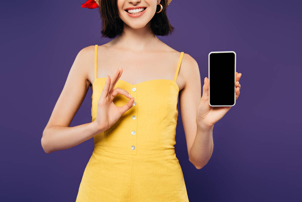 oříznutý pohled na usmívající se dívku v přidržující se smartphone s prázdnou obrazovkou a ukazující na OK nápis izolovaný na purpurovém  - Fotografie, Obrázek