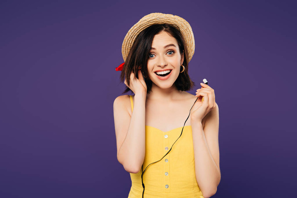 excited smiling pretty girl in straw hat putting on earphones isolated on purple - Photo, image