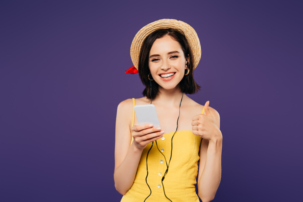 sonriente chica bonita en sombrero de paja escuchando música en los auriculares en el teléfono inteligente y mostrando el pulgar hacia arriba aislado en púrpura
 - Foto, imagen