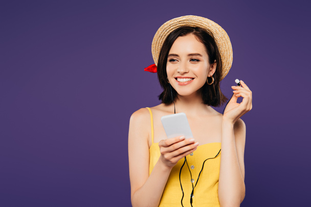 lächelndes hübsches Mädchen mit Strohhut, das Musik in Kopfhörern auf dem Smartphone hört und isoliert auf violettem Grund wegschaut - Foto, Bild
