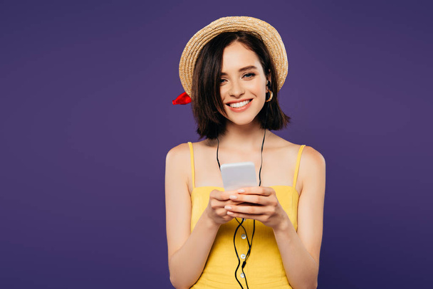 smiling pretty girl in straw hat listening music in earphones on smartphone isolated on purple - 写真・画像