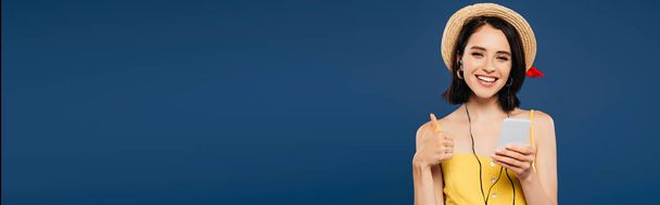 Panoramaaufnahme eines lächelnden hübschen Mädchens mit Strohhut, das Musik über Kopfhörer auf dem Smartphone hört und den Daumen vereinzelt auf blau zeigt - Foto, Bild