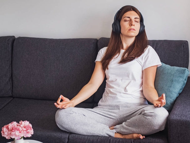 Young woman in big headphones sitting on the sofa and listening to calm music and meditating - Foto, afbeelding