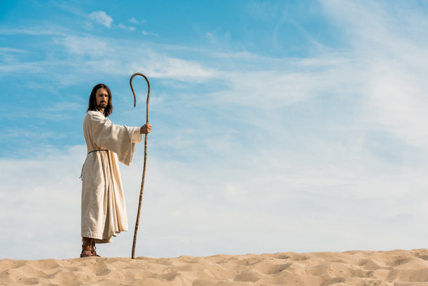 handsome man in jesus robe holding wooden cane against sky in desert  - Foto, immagini