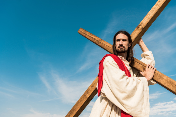 Blick auf den bärtigen Jesus, der ein Kreuz gegen den Himmel mit Wolken hält  - Foto, Bild