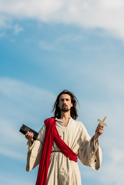 ο Ιησούς κρατώντας ιερή Βίβλο και σταυρό ενάντια στον γαλάζιο ουρανό με σύννεφα  - Φωτογραφία, εικόνα