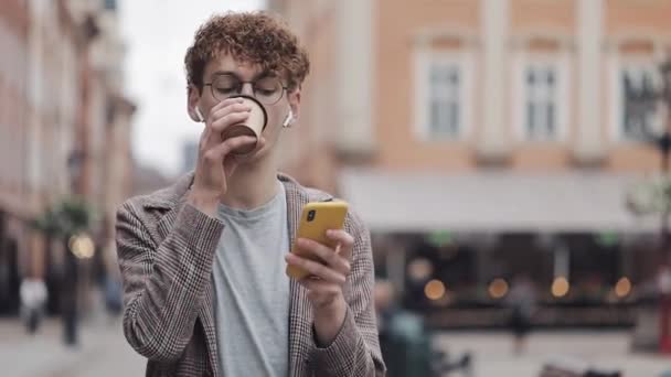Young Hipster Guy in Glasses and with Earphones Wearing Checked Jacket Using His Smartphone Typing Message in Social Networks, Drinking Coffee Walking at Old City Street. Communication Concept. - Video, Çekim