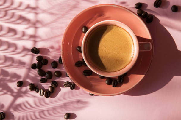 Coral colored cup with effuse coffee's beans with shadows on pastel rose background. Flat lay. Top view. Coffee concept. Pop art still life - Photo, Image
