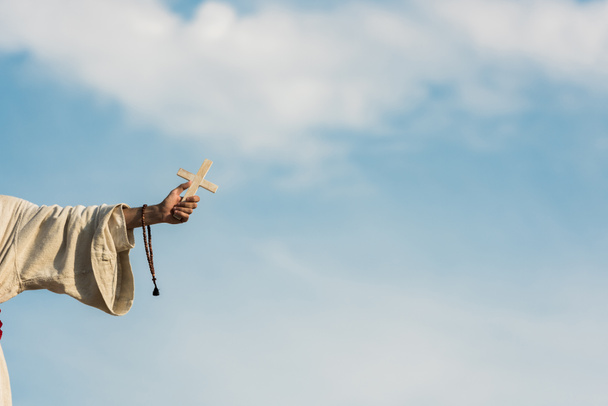 cropped view of religious man holding cross and rosary beads against blue sky  - Foto, Bild