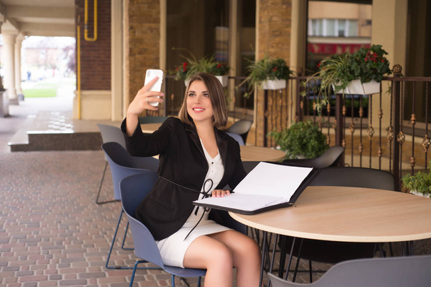 Chica de negocios sentada en el café con la carpeta de documentos, haciendo selfie
 - Foto, Imagen