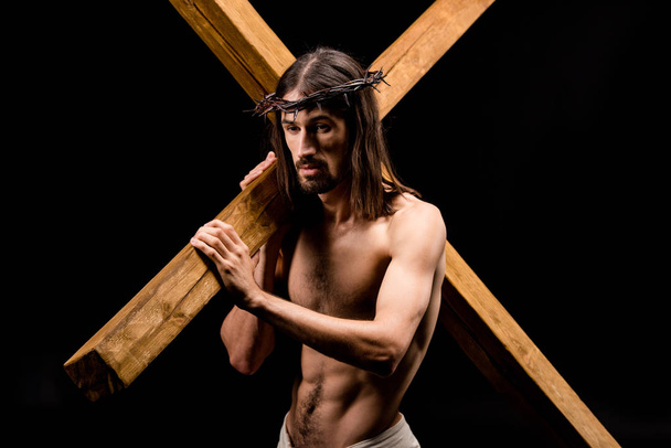 upset shirtless man in wreath holding cross isolated on black  - Фото, зображення