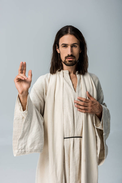 jóképű férfi Jézus köntös kézzel a mellkason a gesztusokkal elszigetelt szürke  - Fotó, kép