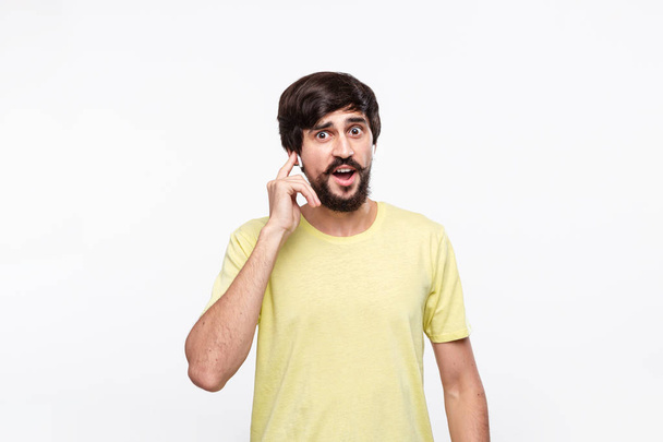 Komea ruskeaverikkö mies keltaisessa paidassa, jossa parta ja mostaatit hymyilevät puhuessaan älypuhelimella, joka seisoo eristettynä valkoisella taustalla. Viestinnän käsite. Hyviä uutisia puhelimitse.
. - Valokuva, kuva