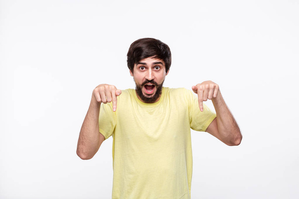 Handsome brunet man in a yellow shirt with beard and mustaches pointing with his fingers down standing isolated over white background. Place of product and advertisment. - Photo, Image