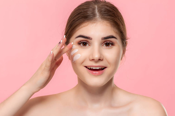 Beautiful model applyes moisturizing cosmetic product to skin holding white tube of a hyaluronic cream isolated over pink background. Concept of beauty and health treatment. - Photo, Image