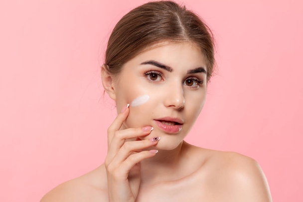 Beautiful model applyes moisturizing cosmetic product to skin holding white tube of a hyaluronic cream isolated over pink background. Concept of beauty and health treatment. - Фото, изображение