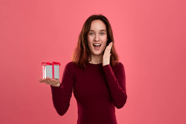 Excited pretty brunette girl holding present box with a ribbon standing isolated on a dark pink background and smiling at the camera. Celebrating special event. - Photo, Image