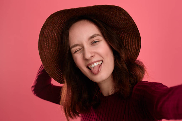 Excited beautiful brunette girl in a wide-brimmed hat making selfie putting out her tongue standing on a dark pink background. - Photo, Image
