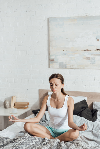 upset young woman sitting on bed in lotus pose with closed eyes - Foto, afbeelding