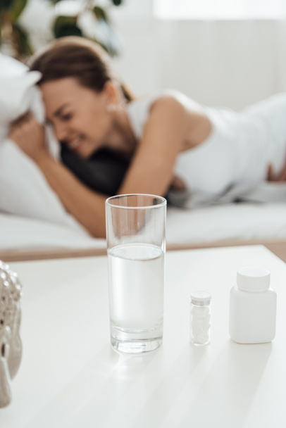 επιλεκτική εστίαση της γυναίκας που υποφέρει στο κρεβάτι και χάπια με ποτήρι νερό στο προσκήνιο - Φωτογραφία, εικόνα