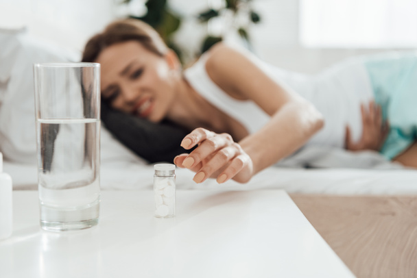 selectieve focus van het lijden van de vrouw in bed en pillen met glas water op de voorgrond - Foto, afbeelding