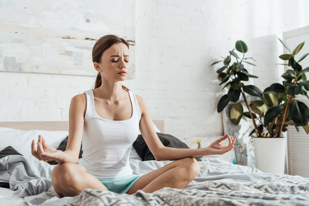 upset young woman sitting on bed in lotus pose with closed eyes - Foto, imagen