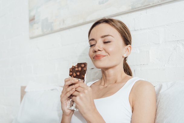 lage hoek uitzicht van lachende jonge vrouw eten chocolade met noten met gesloten ogen in bed - Foto, afbeelding