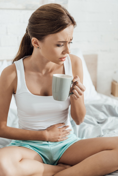 stresli genç kadın yatakta oturan, göbek dokunmadan ve çay fincan tutarak - Fotoğraf, Görsel